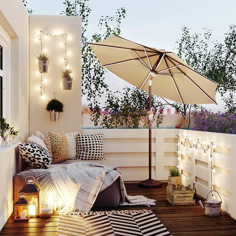 Patioslife Solara tan market umbrella on a cosy balcony lounge with warm outdoor lights and garden seating