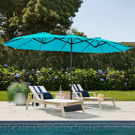 Patioslife Horizon Haven double-sided patio umbrella with base in sky blue, 4.6 x 2.7m,with LED lights over two loungers beside a pool with garden plants